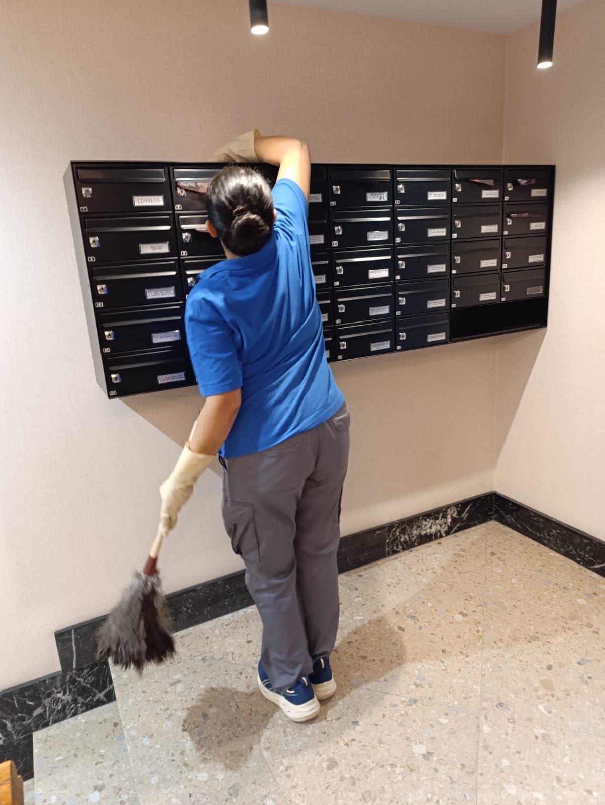 Mujer con camisa azul quitando el polvo de los buzones de correo en un pasillo.