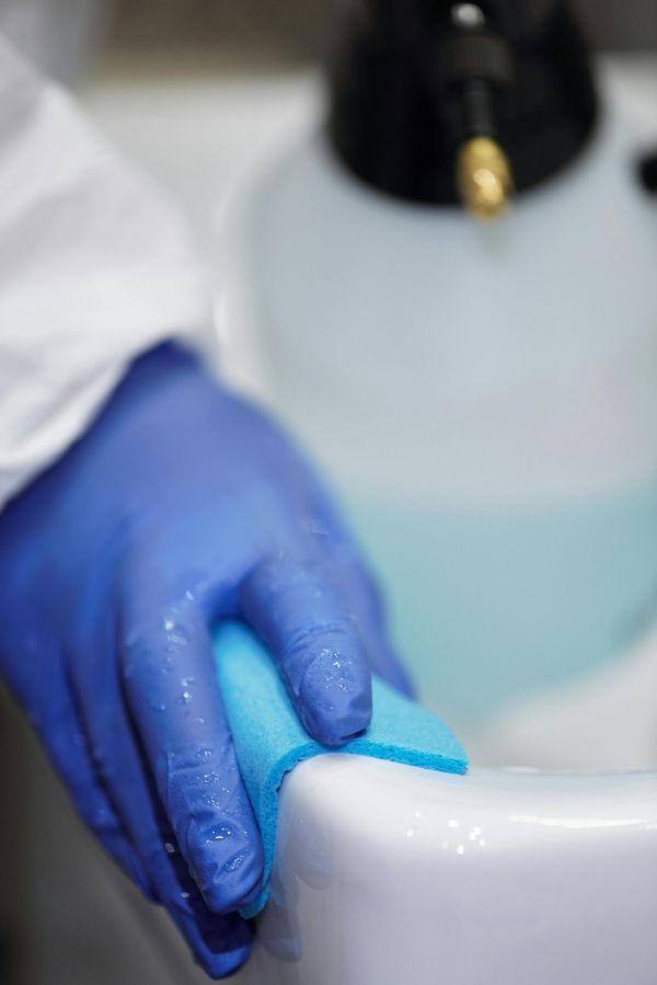 Persona con guantes azules limpiando una superficie blanca con una esponja azul, con un pulverizador desinfectante al fondo.