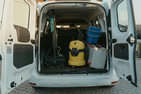 La parte trasera abierta de la camioneta blanca revela equipos de limpieza: una aspiradora, un balde y una hidrolavadora, listos para trabajar.