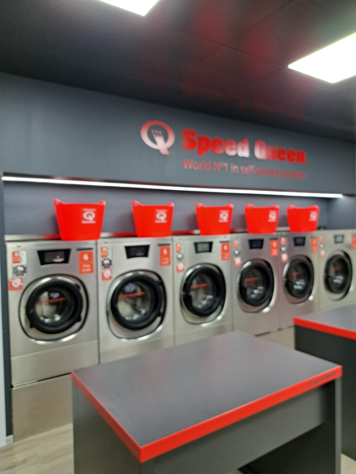 Una hilera de lavadoras Speed ​​Queen de acero inoxidable, coronadas con cestas de ropa rojas a juego, en una lavandería moderna.