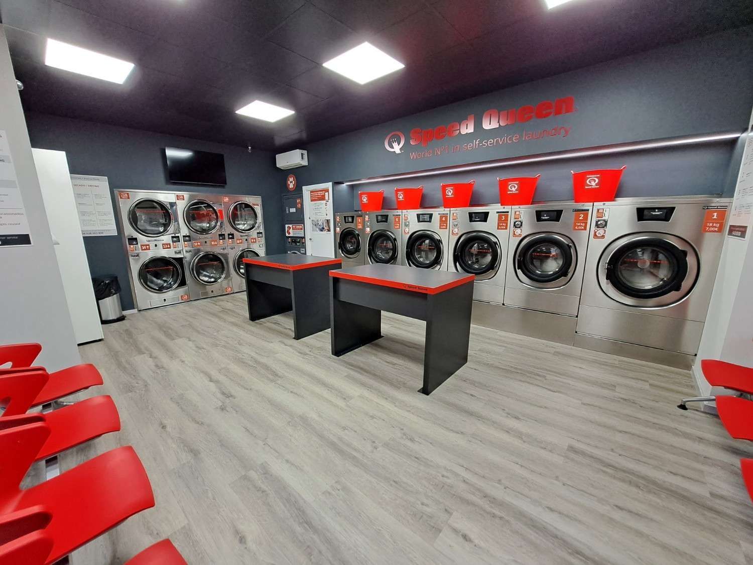 Una lavandería moderna y limpia con filas de lavadoras Speed ​​Queen de acero inoxidable, dos mesas plegables y asientos rojos.