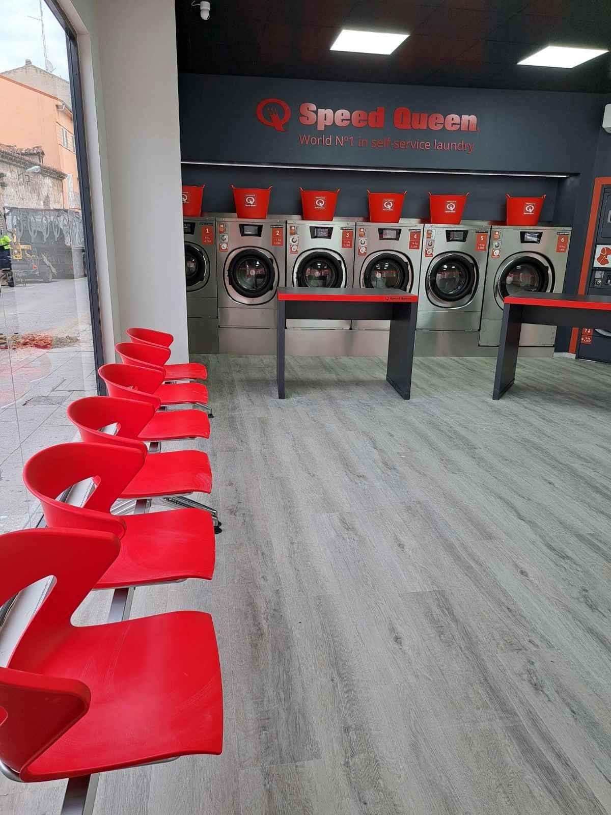 Una hilera de sillas rojas se sitúa frente a una fila de lavadoras en el interior de una lavandería moderna con suelos de imitación de madera gris.