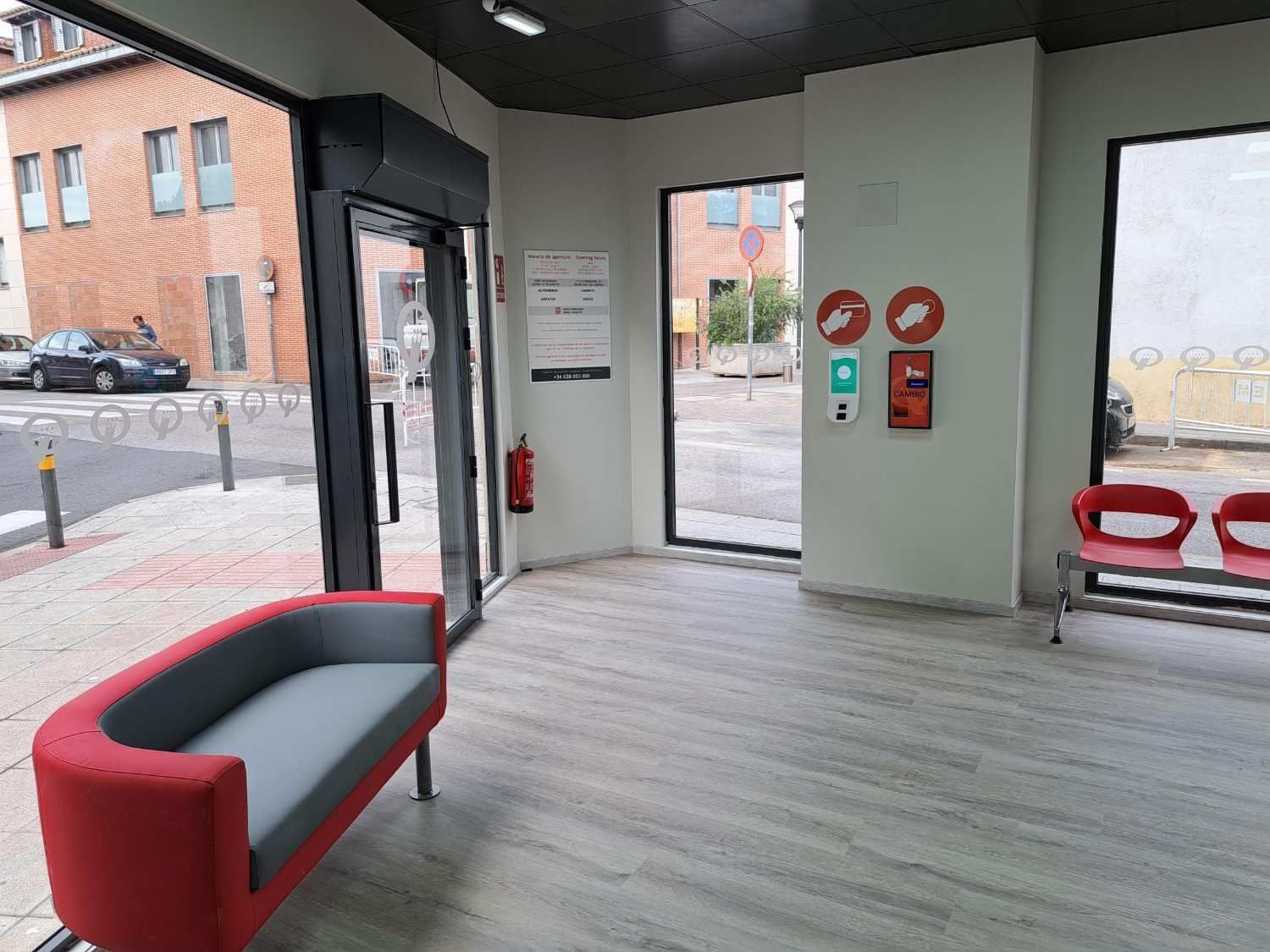 Una sala de espera interior con un sofá curvo rojo y gris, sillas rojas, puertas de cristal y señalización de seguridad en paredes blancas.