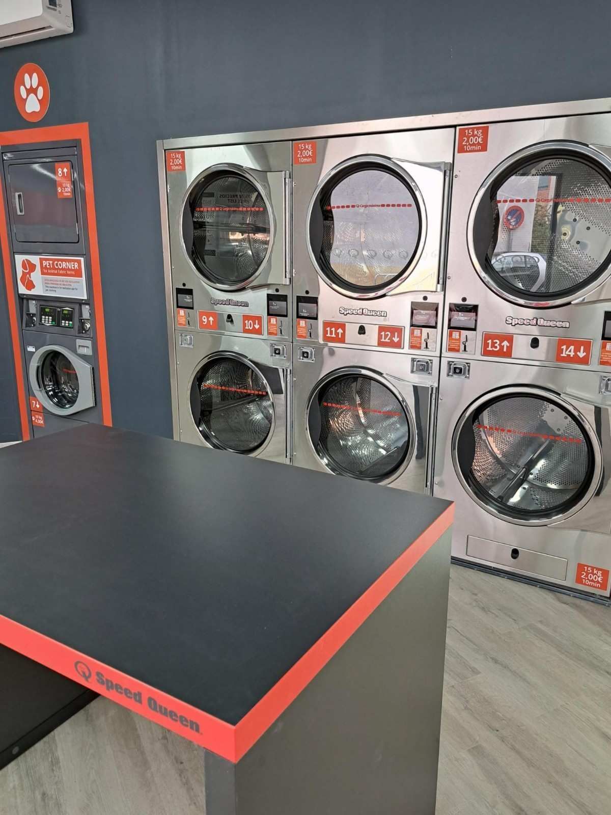 Una hilera de lavadoras industriales apiladas de acero inoxidable y una mesa para doblar la ropa dentro de una lavandería limpia de paredes grises.