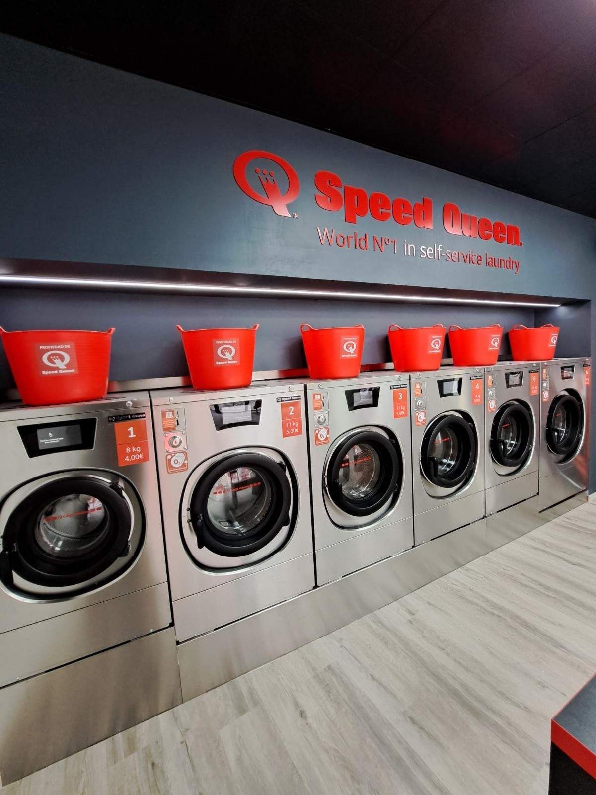 Una hilera de lavadoras Speed ​​Queen de acero inoxidable en una lavandería, cada una coronada con una cesta de ropa roja a juego.