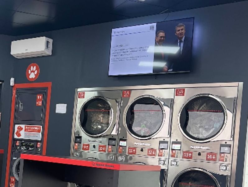 Una lavandería con un televisor montado en la pared que muestra una presentación sobre tres secadoras de acero inoxidable.