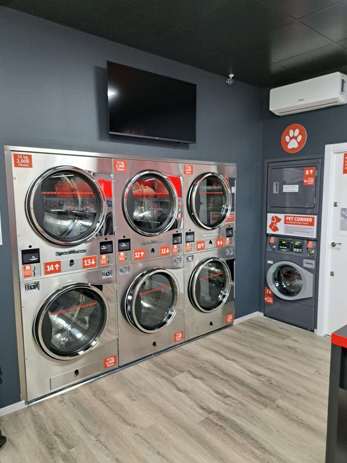 Una hilera de secadoras de acero inoxidable con una zona de lavado exclusiva para mascotas y un televisor en una pared gris oscuro en un luminoso cuarto de lavandería.