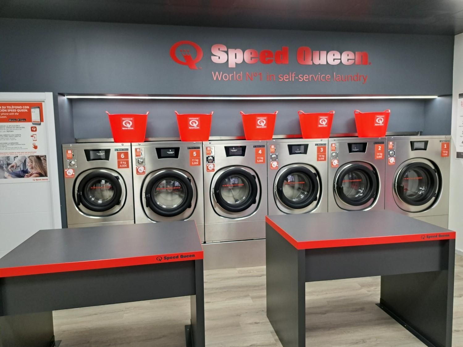 Una hilera de seis lavadoras Speed ​​Queen de acero inoxidable con cestas de ropa rojas encima en un cuarto de lavado.