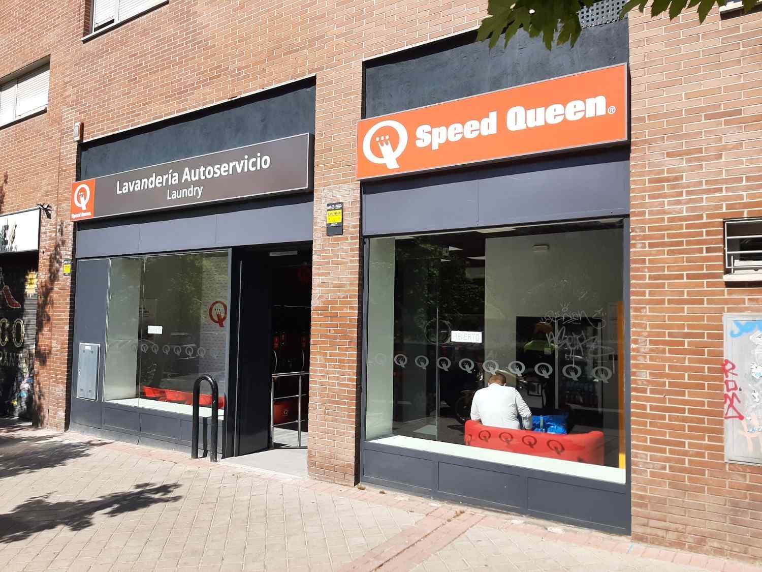 Una lavandería Speed ​​Queen con paredes de ladrillo, molduras grises y una persona sentada en un banco rojo dentro del escaparate de cristal.