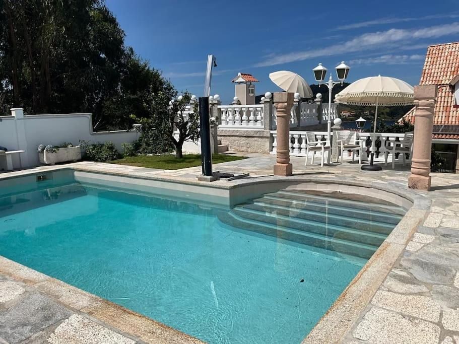 Piscina con escaleras, patio de piedra, columnas, sombrillas y un edificio en un día soleado.