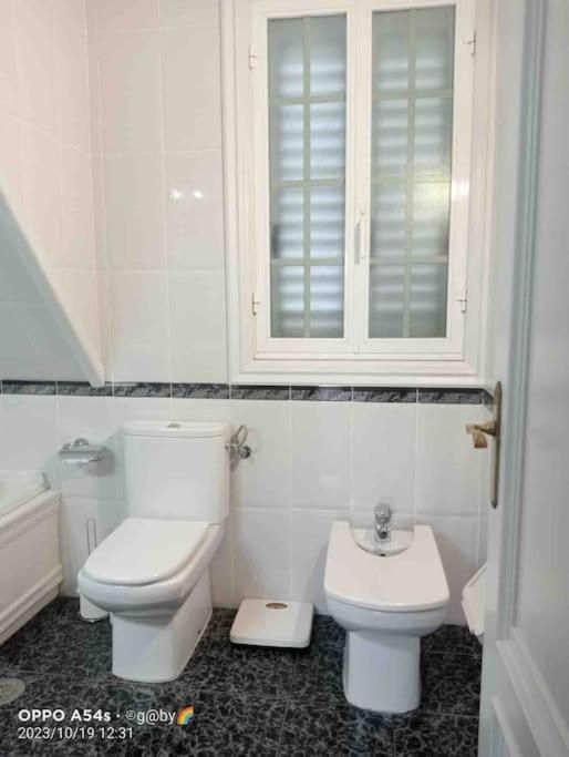 Baño con inodoro y bidet blancos, suelo de mármol y ventana con persianas blancas.