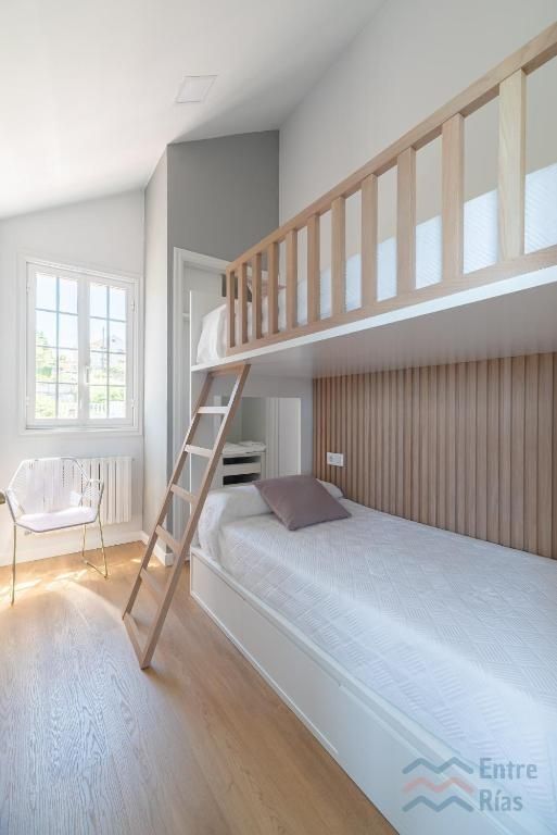 Litera en una habitación luminosa con suelo de madera clara y escalera de madera. Ropa de cama blanca y silla.