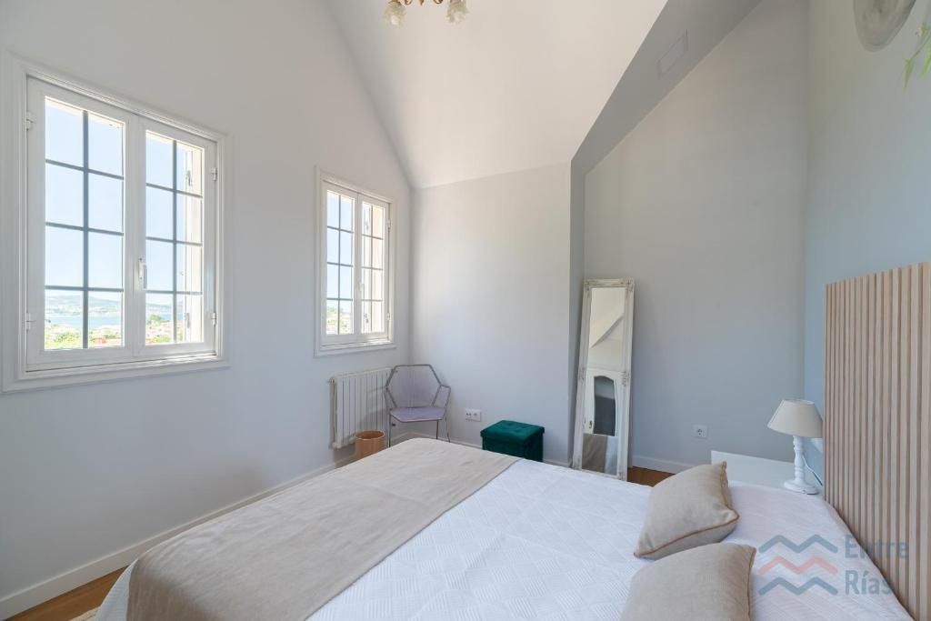 Dormitorio con cama, ventanas y espejo de cuerpo entero. Paredes blancas y luz natural.