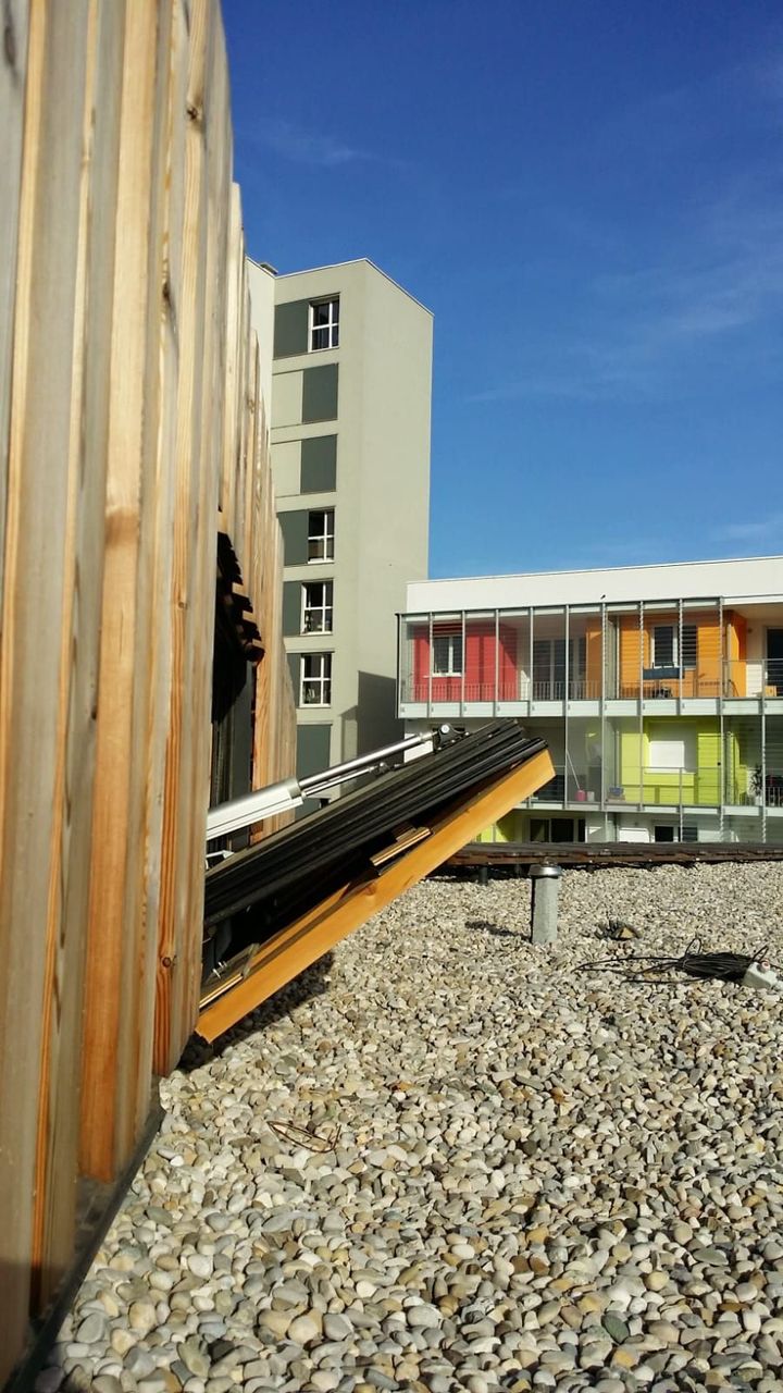 Façade d'un bâtiment en bois à côté d'une terrasse en gravier sur le toit. Bâtiments modernes en arrière-plan sous un ciel bleu.