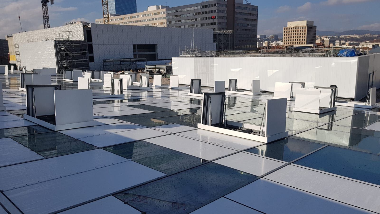 Vue d'un toit avec un motif de tuiles de verre et des caissons de ventilation blancs, avec des bâtiments en arrière-plan. Journée ensoleillée.