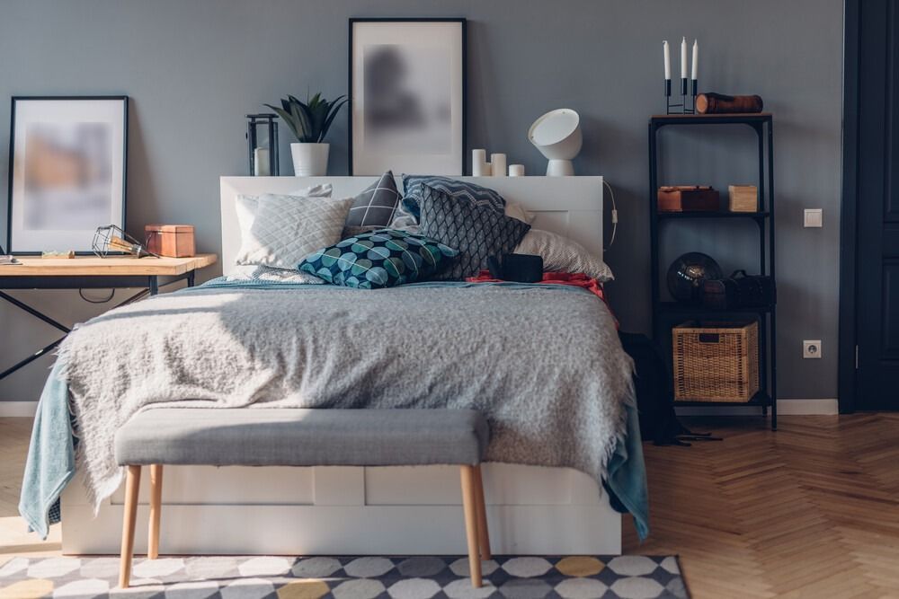 Dormitorio con cama, escritorio y estantería contra una pared gris. La cama tiene varias almohadas y una manta gris claro.