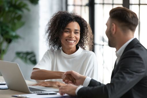 Mujer y hombre se dan la mano en un escritorio, sonriendo. Portátil visible; entorno de oficina.