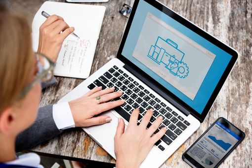Persona trabajando con una laptop, con un notebook y un teléfono cerca. La pantalla muestra un ícono empresarial azul.