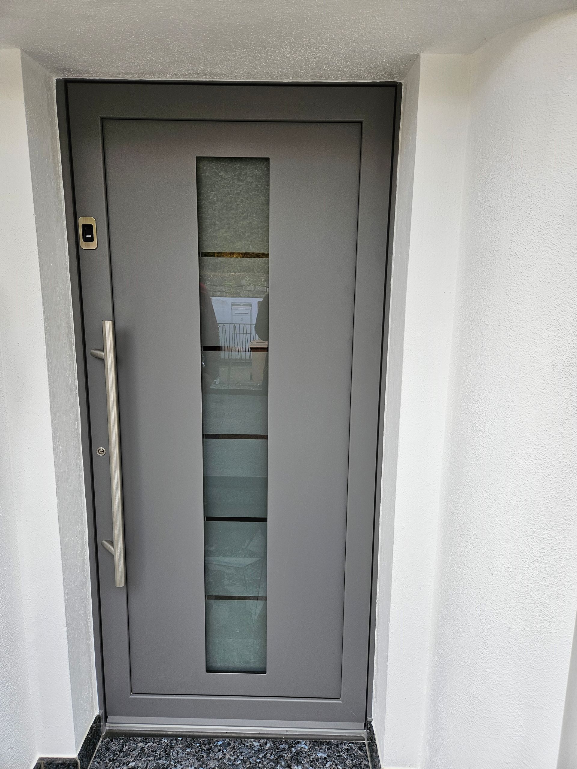 Graue Haustür mit vertikaler Glasscheibe, langem Griff, eingelassen in eine weiße Wand.