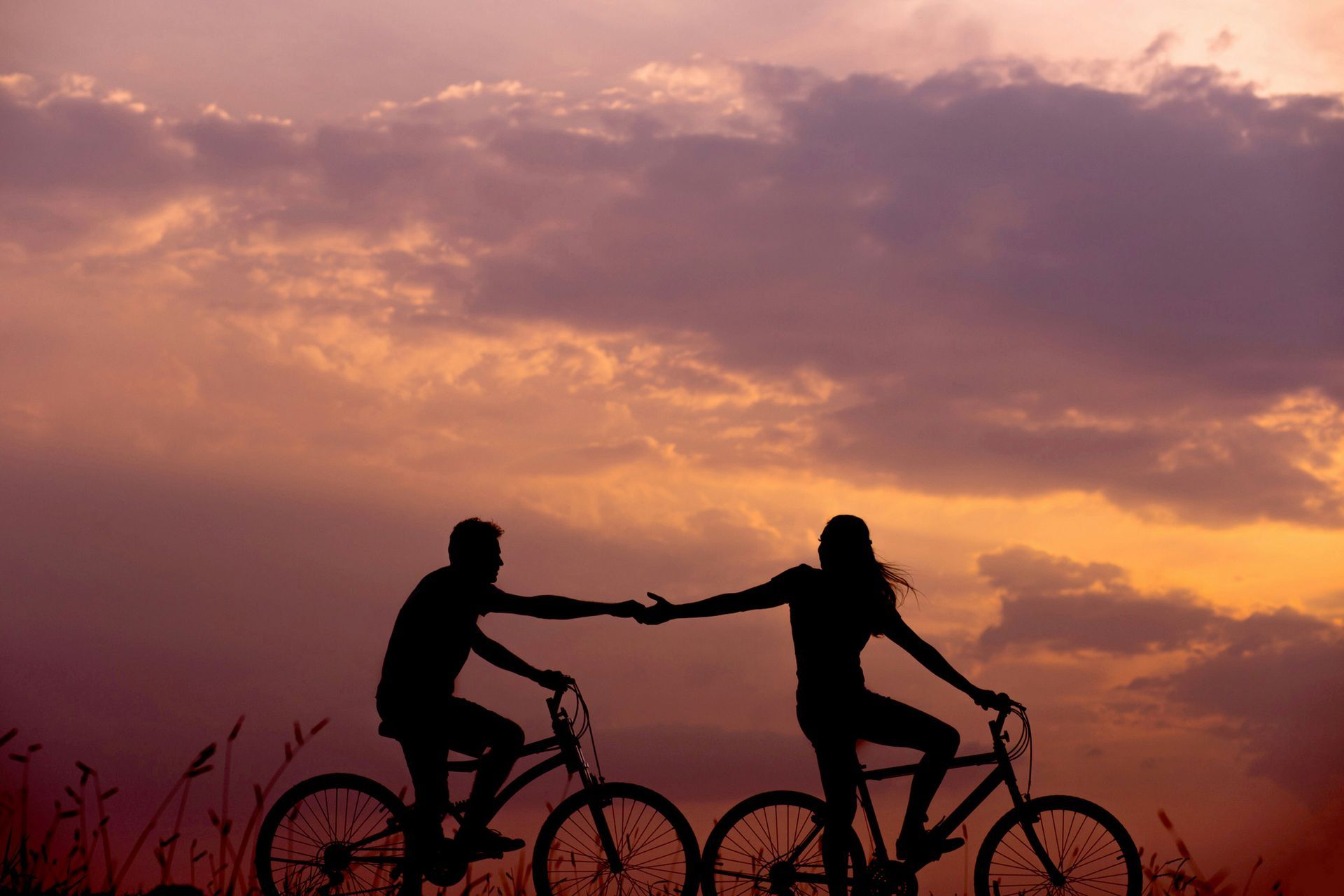 Dos ciclistas recortados contra el cielo al atardecer, extendiendo los brazos el uno hacia el otro en una carretera bajo un cielo rosa anaranjado.