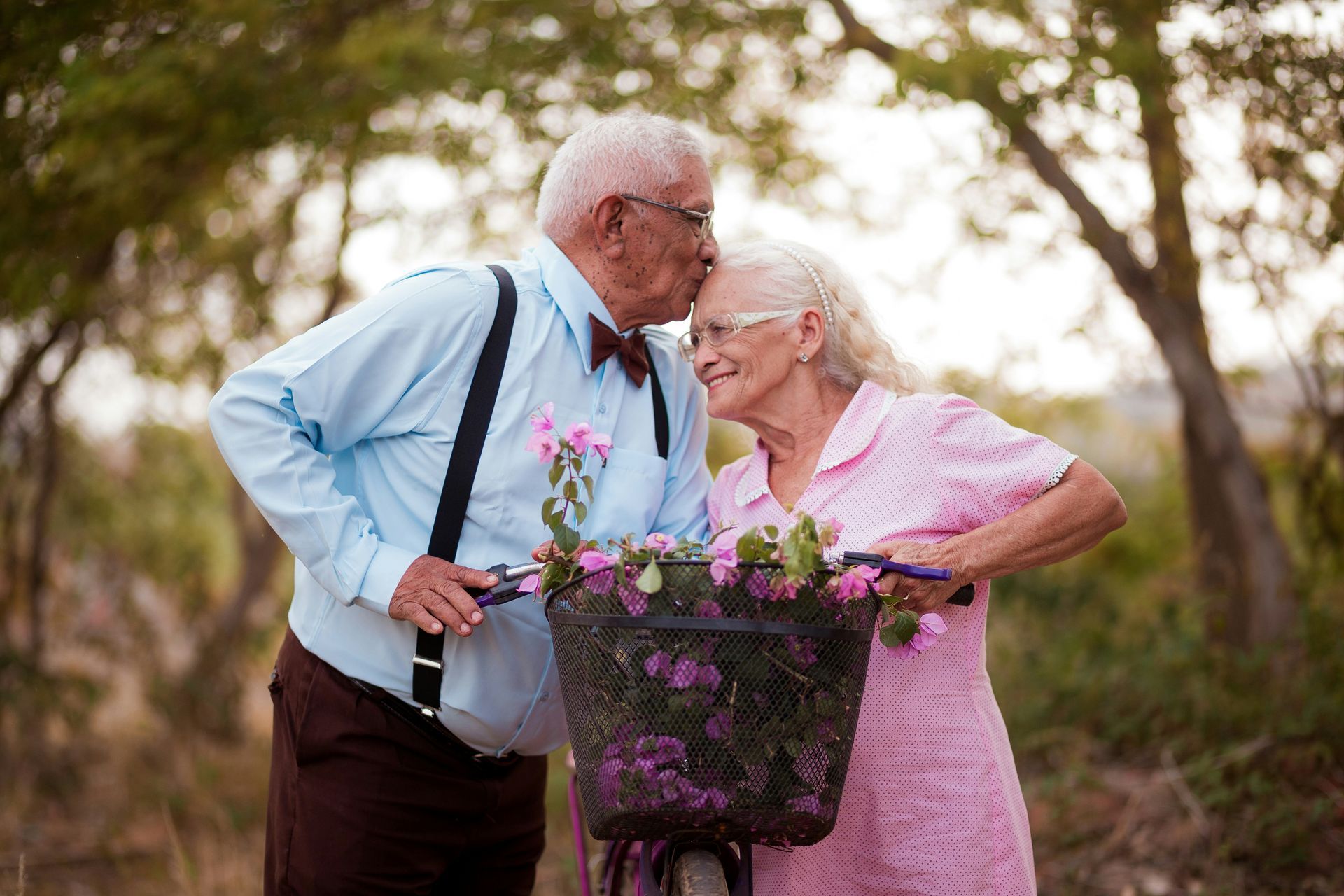 Una pareja sonriente vestida de etiqueta se inclina muy cerca de una bicicleta llena de flores al aire libre.