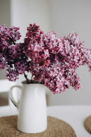 Lilas rosadas en una jarra blanca sobre una estera tejida contra una pared clara.