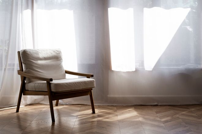 Silla blanca acolchada junto a una ventana soleada con cortinas transparentes en una habitación luminosa.