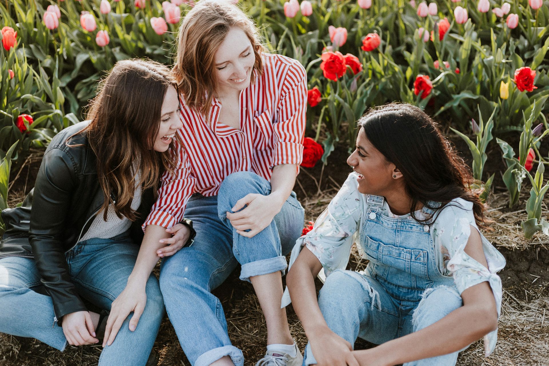Tres amigas sentadas en un campo de tulipanes, riendo y charlando con ropa informal.