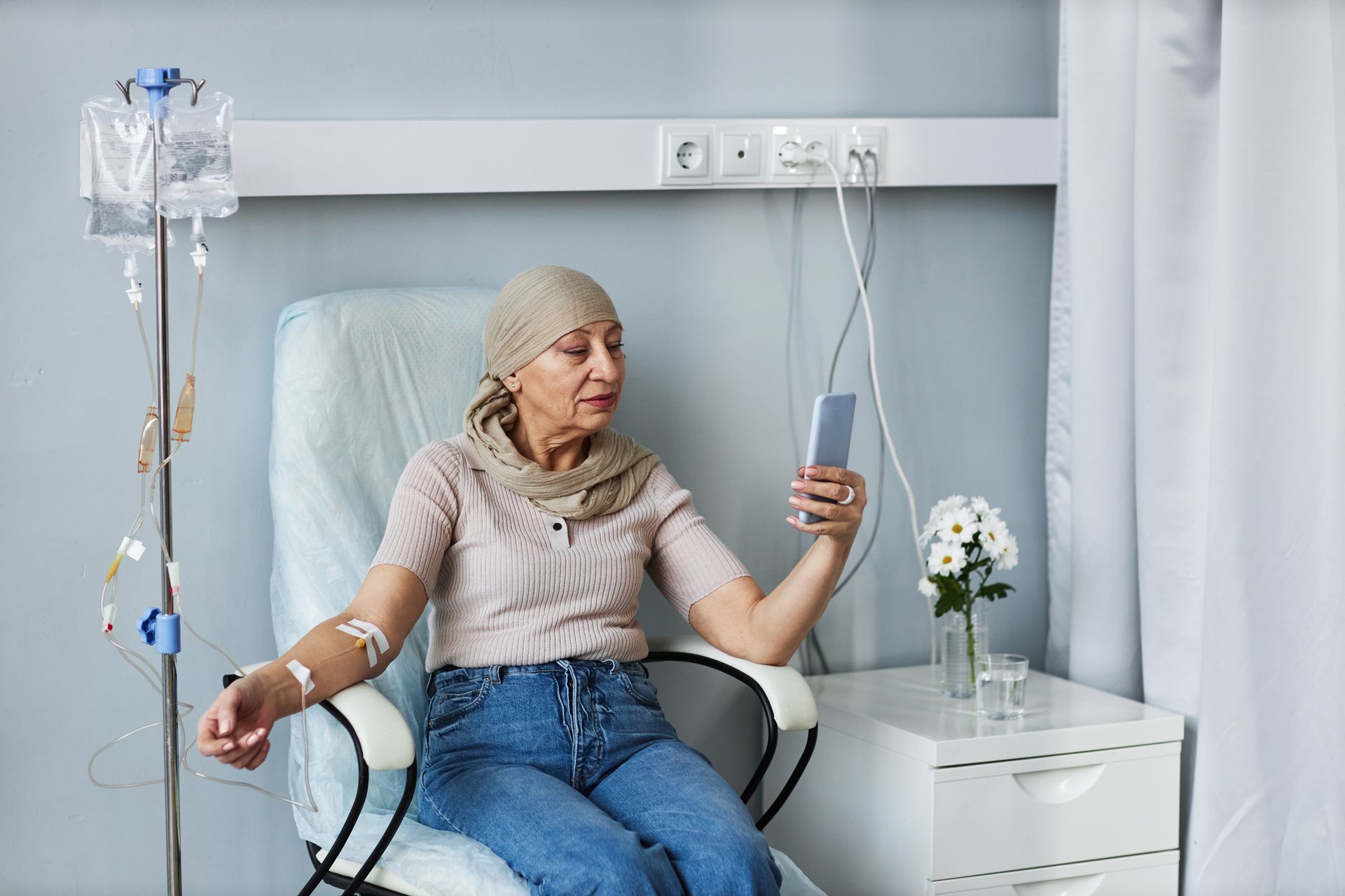Paciente sentado en una silla de hospital con una bolsa de suero intravenoso, con un soporte para el goteo y flores blancas cerca.