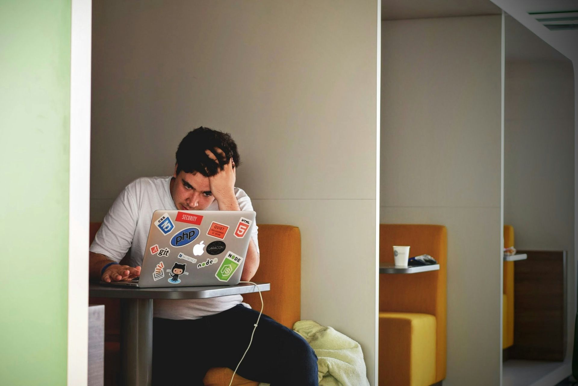 Una persona estudia en una computadora portátil sentada en una mesa en una habitación pequeña, con expresión de frustración y la cabeza entre las manos.