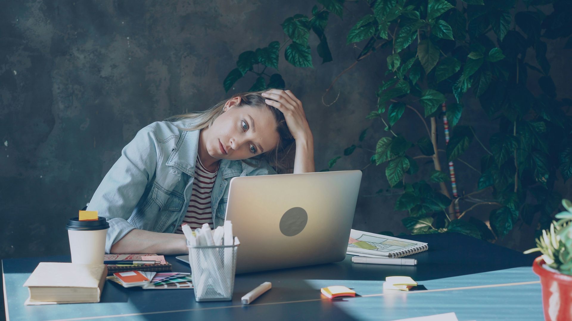 Una persona cansada apoya la cabeza en la mano en un escritorio, mirando una computadora portátil en un espacio de trabajo con poca luz, con papeles y una taza de café.