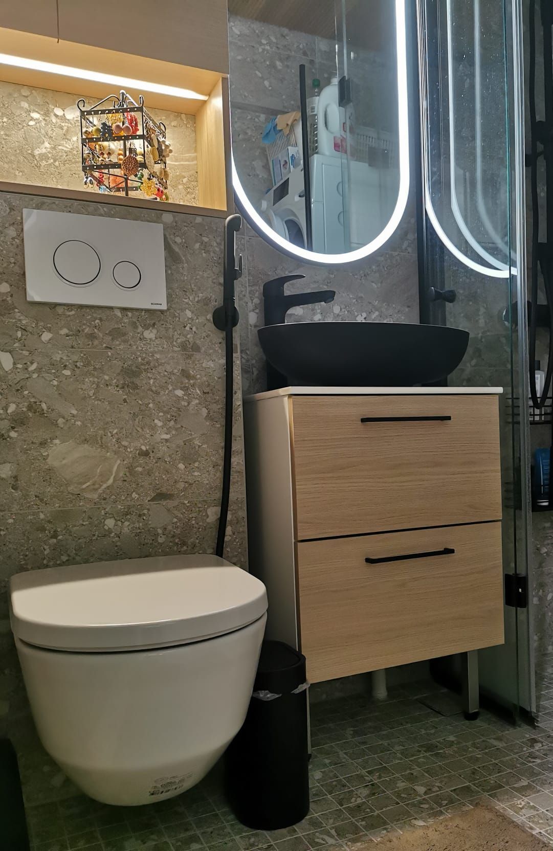 Tyylikäs ja moderni wc-tila, jossa on kivipintainen seinä, musta malja-allas, puunsävyinen allaskaappi ja valaistu peili. Toimiva kokonaisuus syntyy huolellisesta suunnittelusta ja viimeistellystä toteutuksesta.
