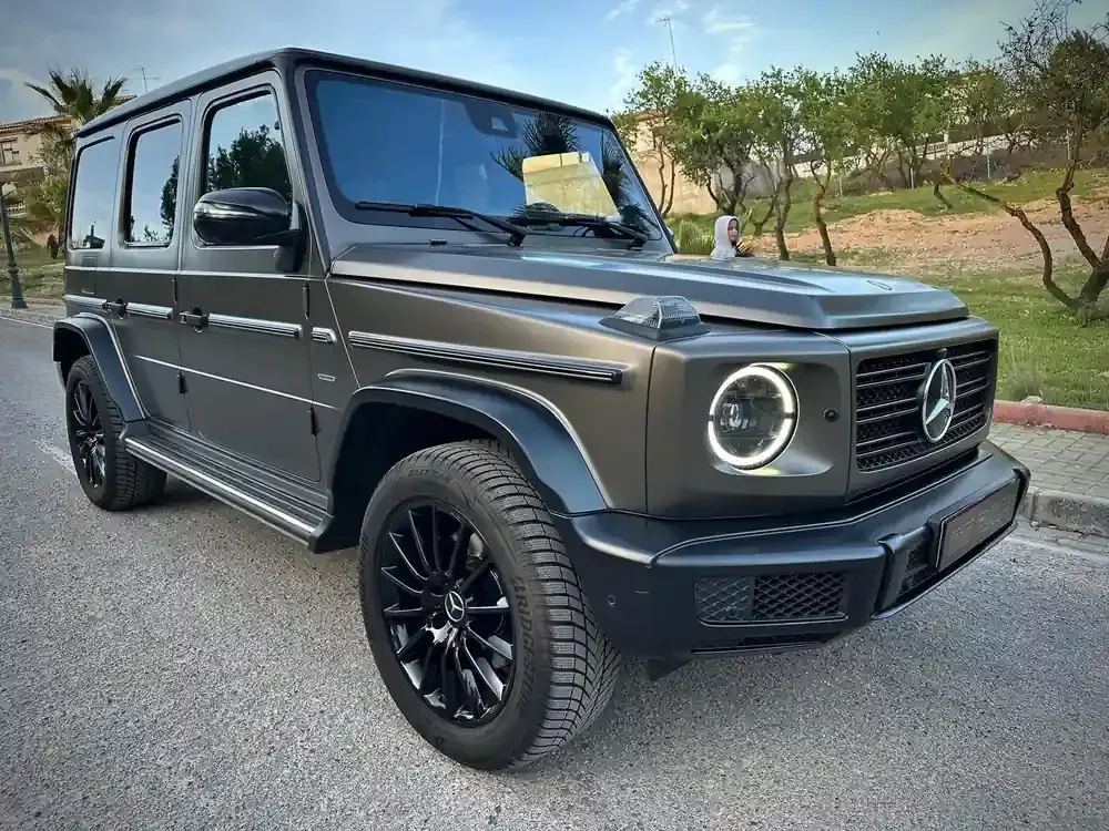 Un SUV Mercedes Clase G de color gris oscuro estacionado en una carretera con árboles al fondo.