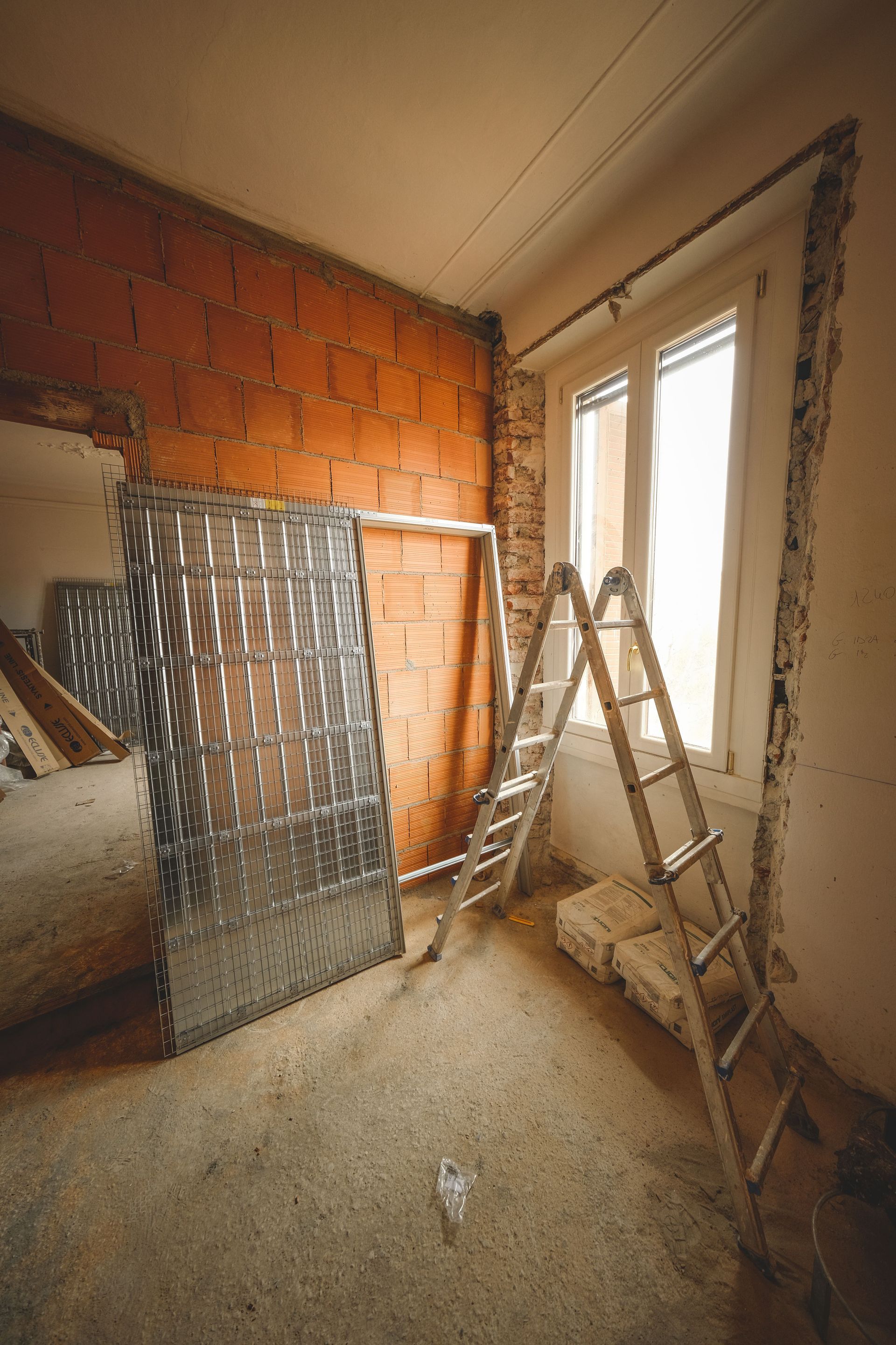 Petite pièce inachevée avec un mur de briques, une échelle, une fenêtre et des matériaux de construction au sol.