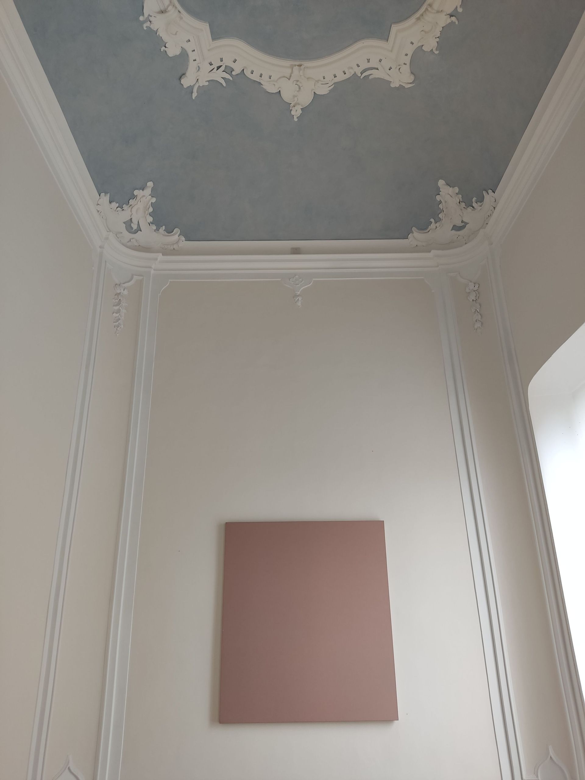 Petite toile rose fixée sur un mur blanc, sous un angle de plafond orné de motifs bleus et blancs.
