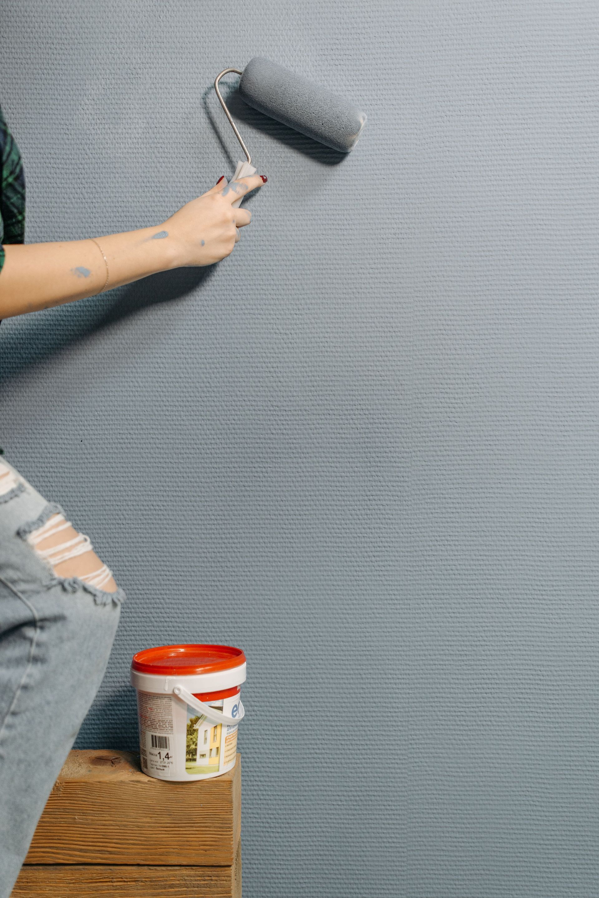 Une personne peint un mur gris texturé au rouleau ; un seau de peinture et un jean déchiré sont visibles.