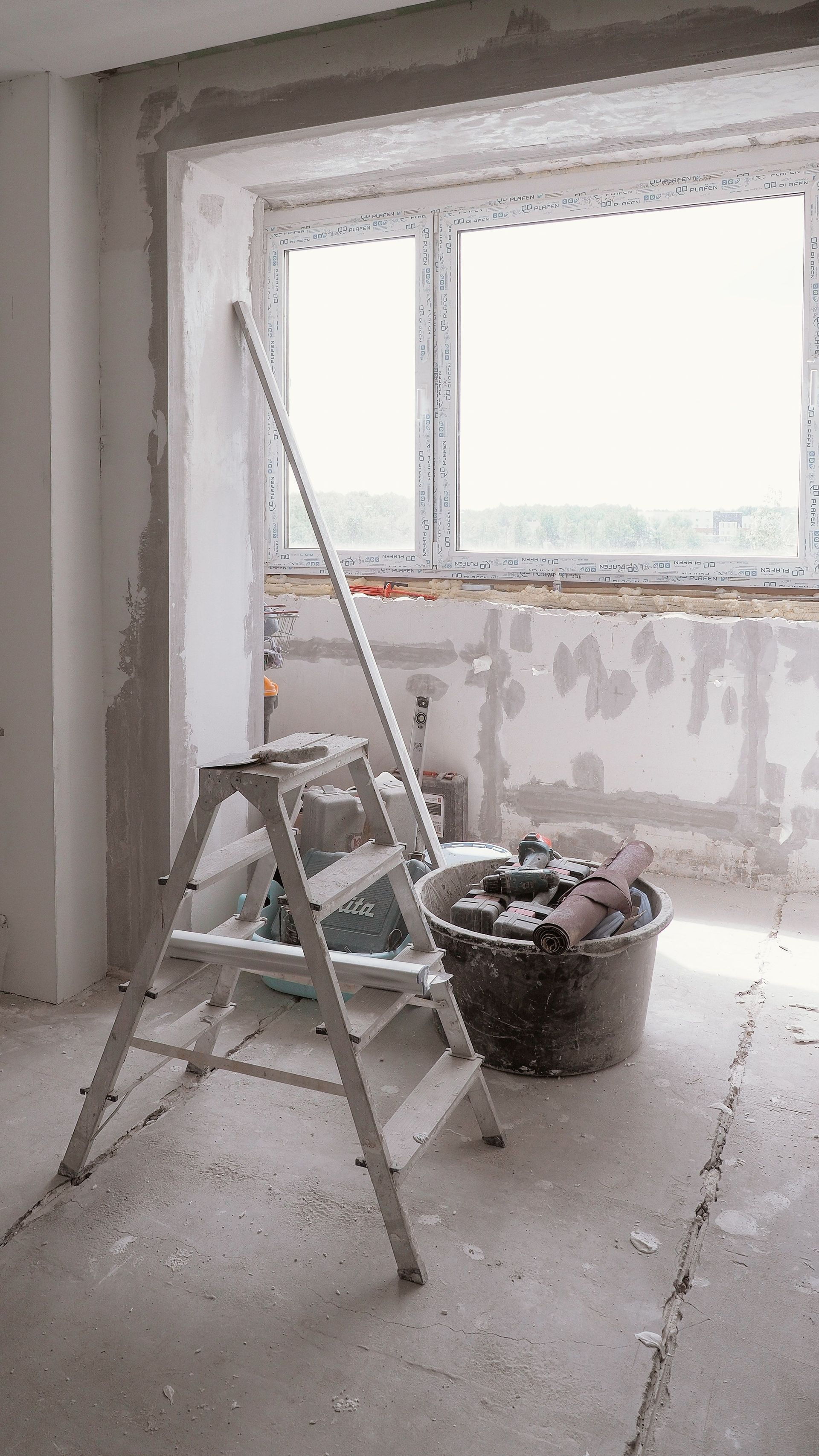 Pièce en rénovation avec escabeau, seau d'outils et mur près de la fenêtre inachevé