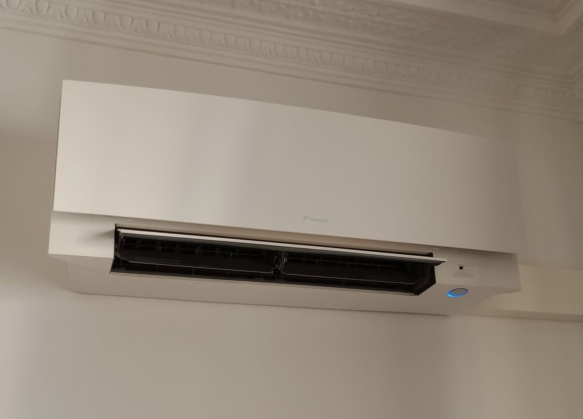 Unité de climatisation blanche montée sur un mur blanc, sous une moulure décorative au plafond.