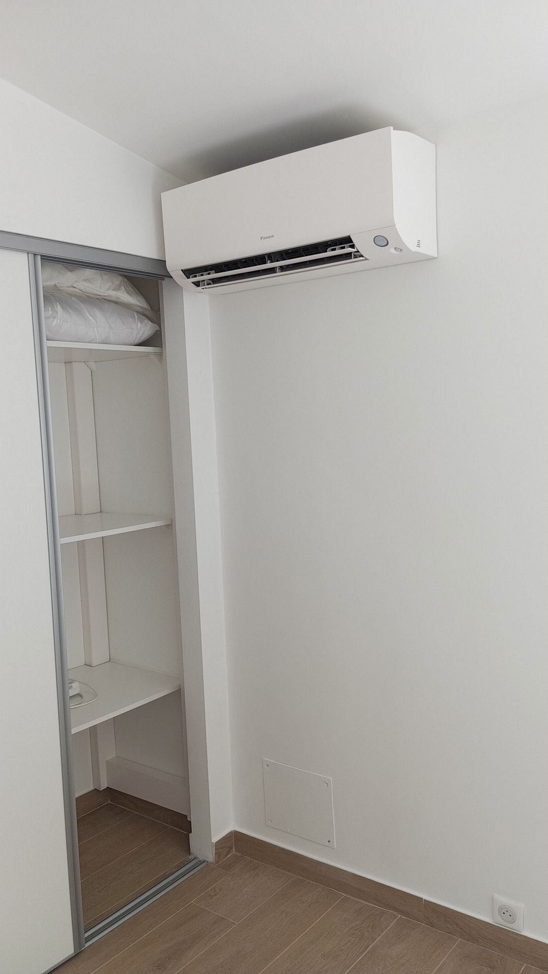 Un climatiseur blanc au-dessus d'un placard à portes coulissantes dans une pièce aux murs blancs et au parquet.