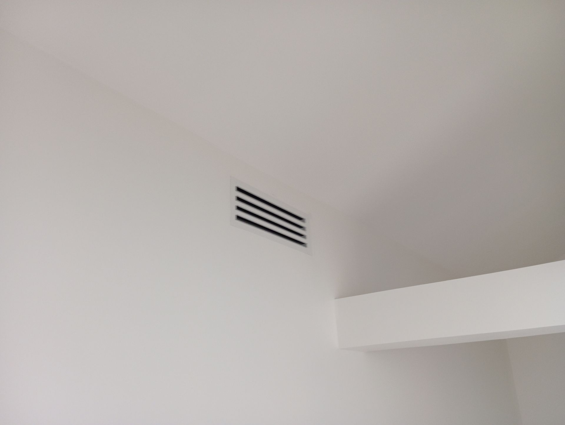 Grille d'aération noire sur un mur blanc, près d'un plafond et d'une poutre blancs.
