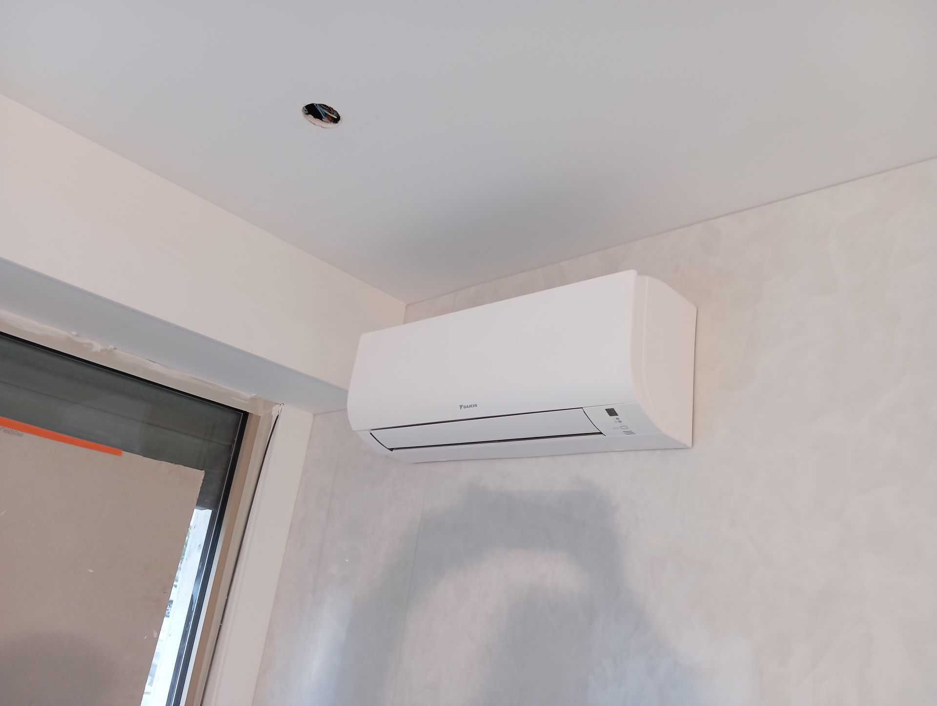 Unité de climatisation blanche montée sur un mur de couleur claire dans une pièce avec une fenêtre.