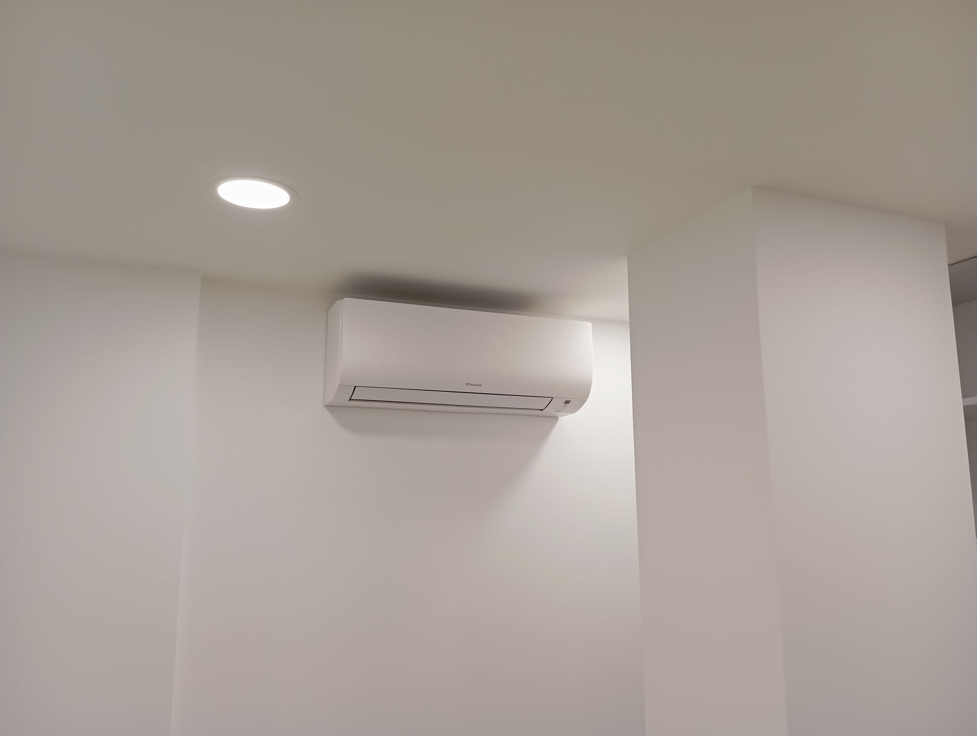 Climatiseur blanc monté sur un mur blanc, sous un plafonnier encastré.