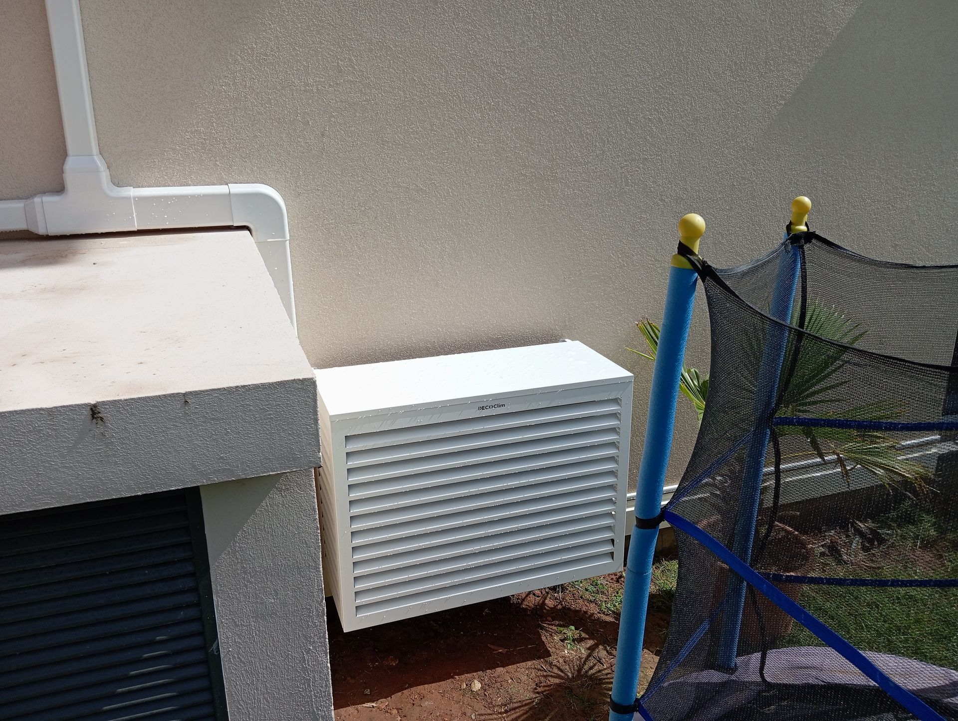 Unité de climatisation blanche montée sur un mur en stuc beige à côté d'un trampoline.
