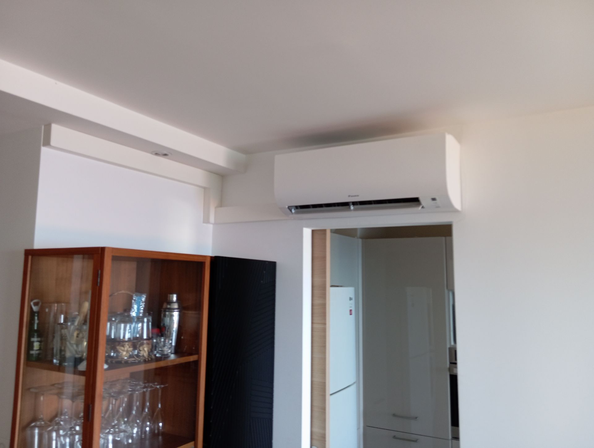 Climatiseur monté sur un mur blanc au-dessus d'une porte et d'une armoire avec verrerie.