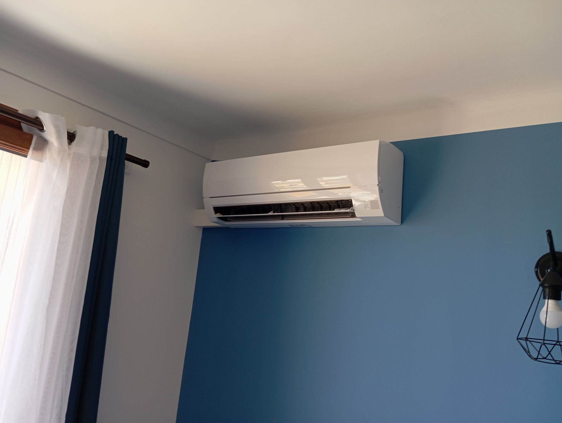 Climatiseur blanc monté sur un mur bleu dans une pièce, à côté d'une fenêtre avec des rideaux.