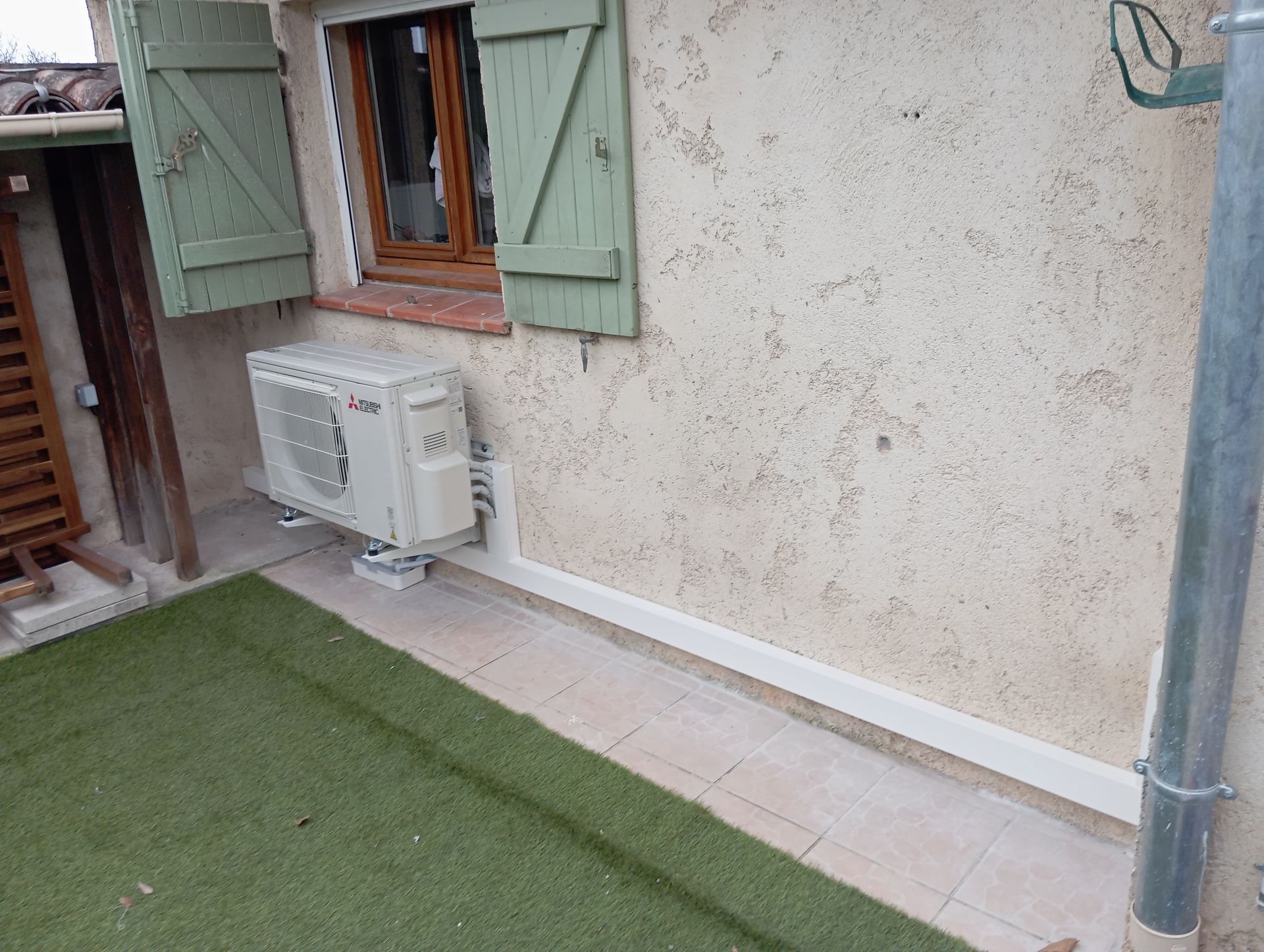 Unité de climatisation sur le mur extérieur d'un bâtiment, près d'une fenêtre et de l'herbe, à côté d'une gouttière.