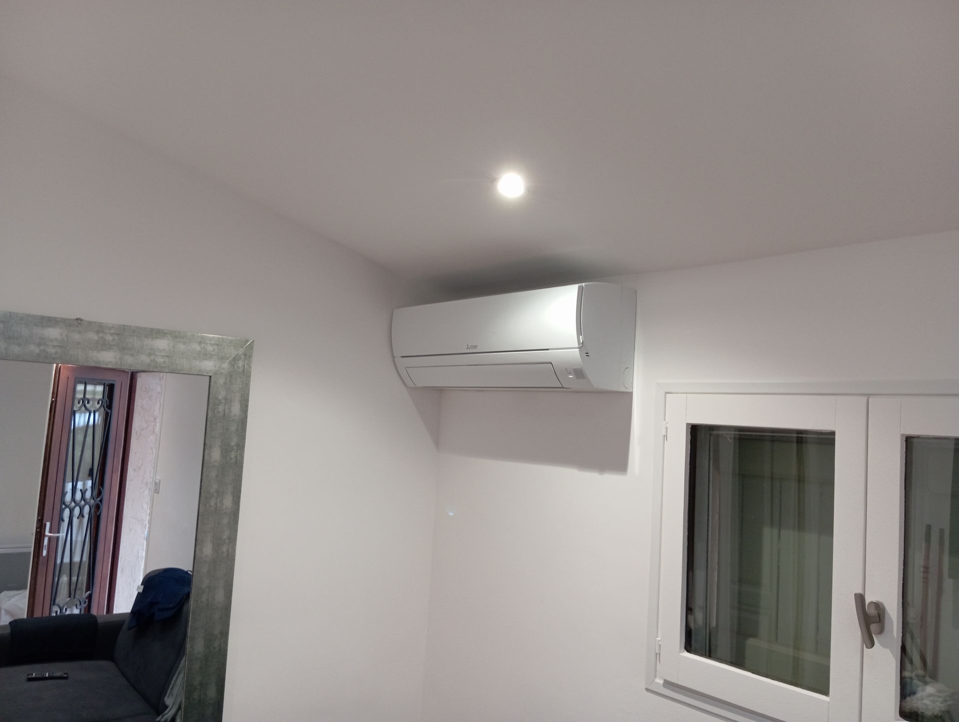 Climatiseur blanc fixé sur un mur blanc, au-dessus d'une fenêtre. On aperçoit un miroir et un canapé.