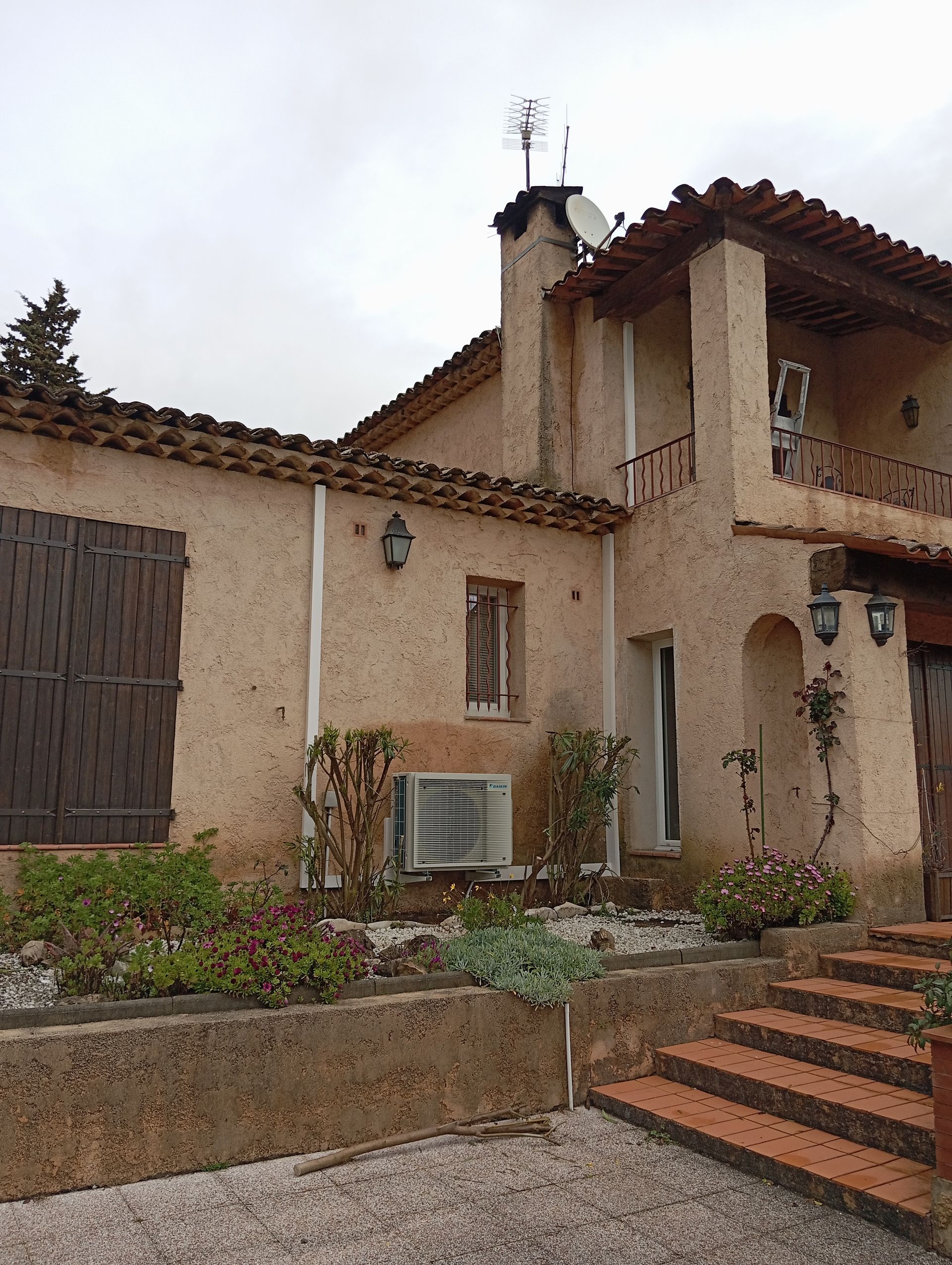 Maison en stuc beige avec toit en tuiles rouges, volets marron et marches menant à l'entrée.