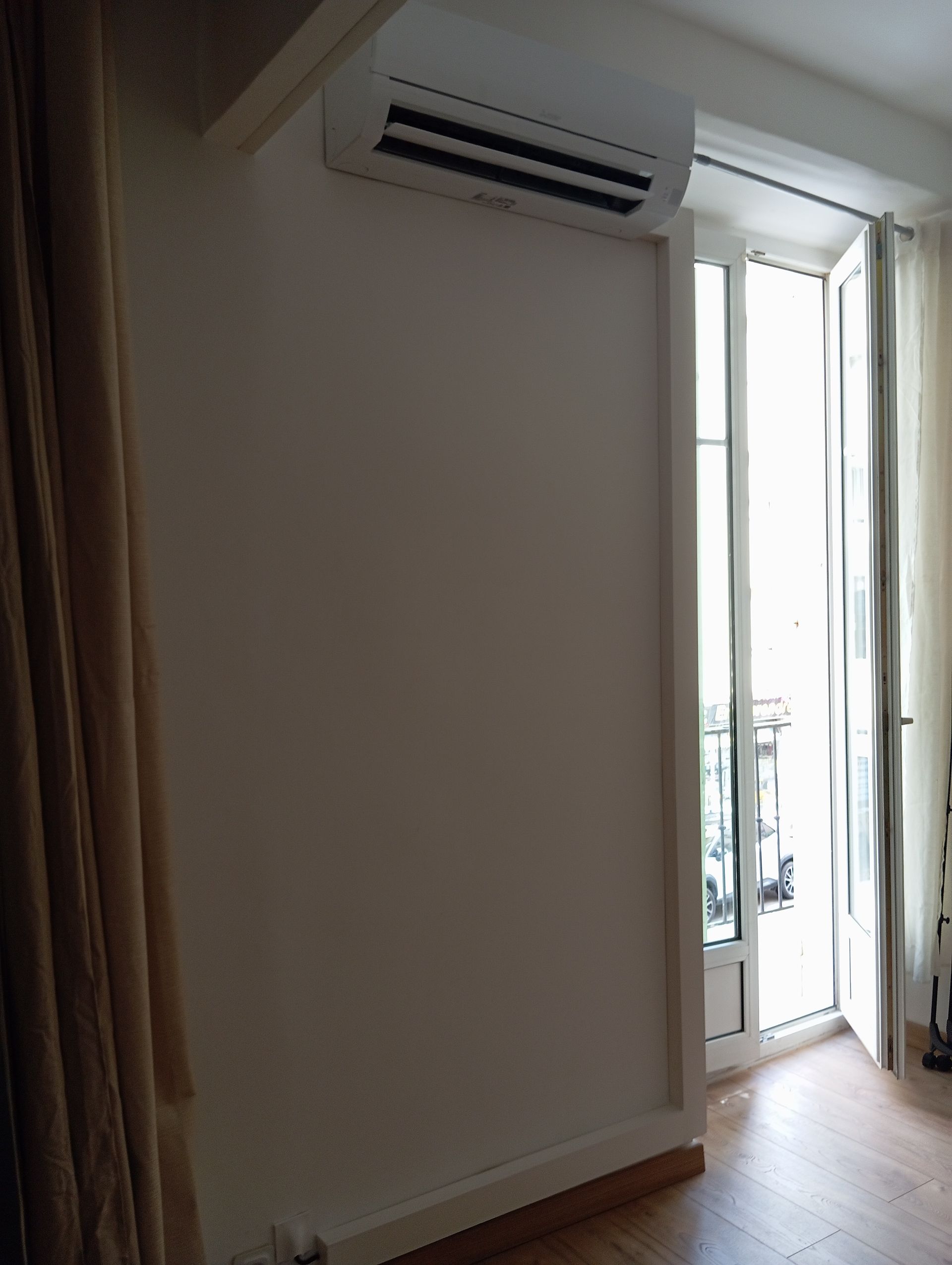 Climatiseur au-dessus d'un mur de couleur crème, à côté d'une fenêtre lumineuse avec des portes ouvertes.