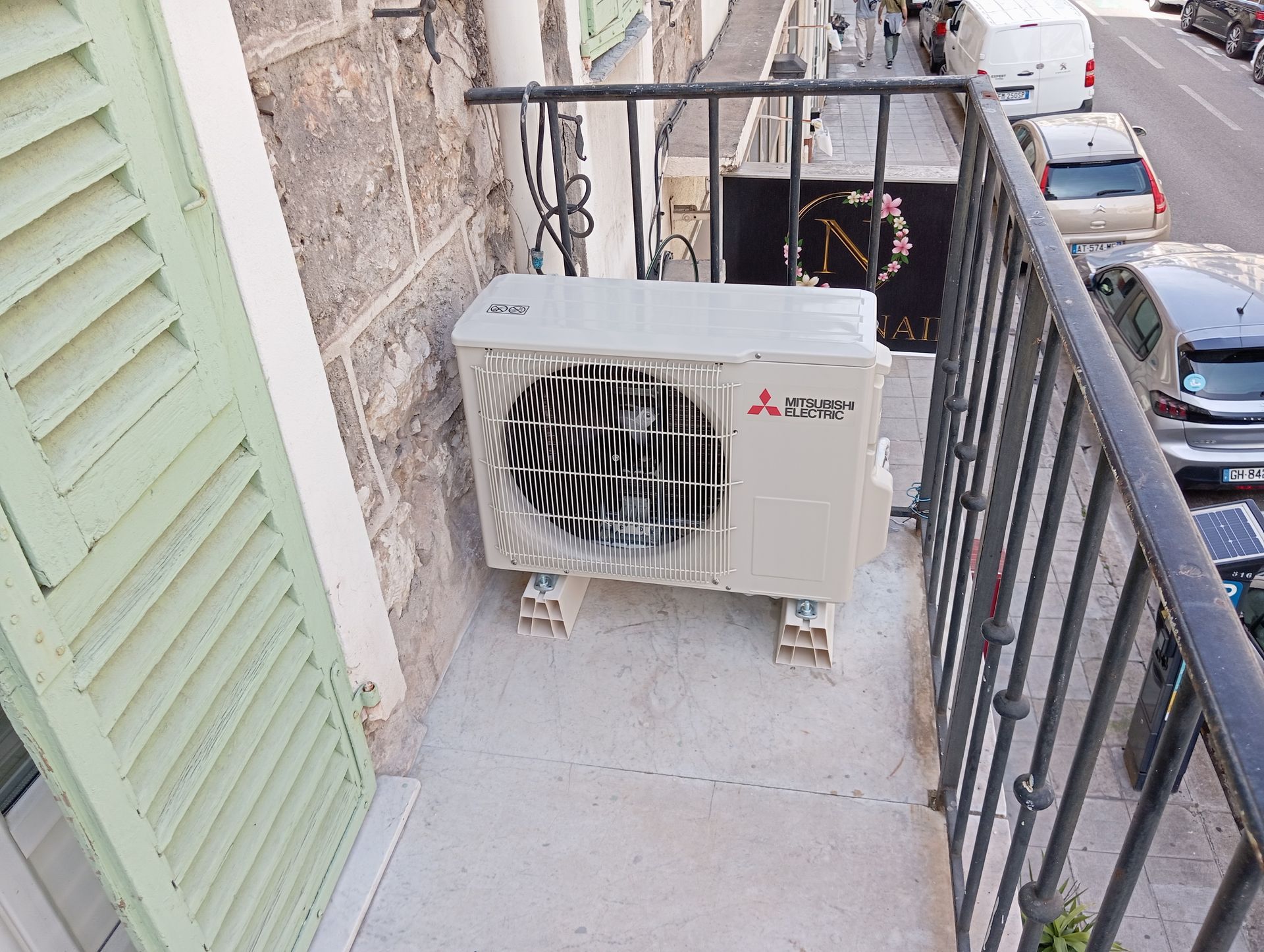 Unité de climatisation extérieure sur un petit balcon avec une balustrade en fer forgé, à côté d'un volet vert.